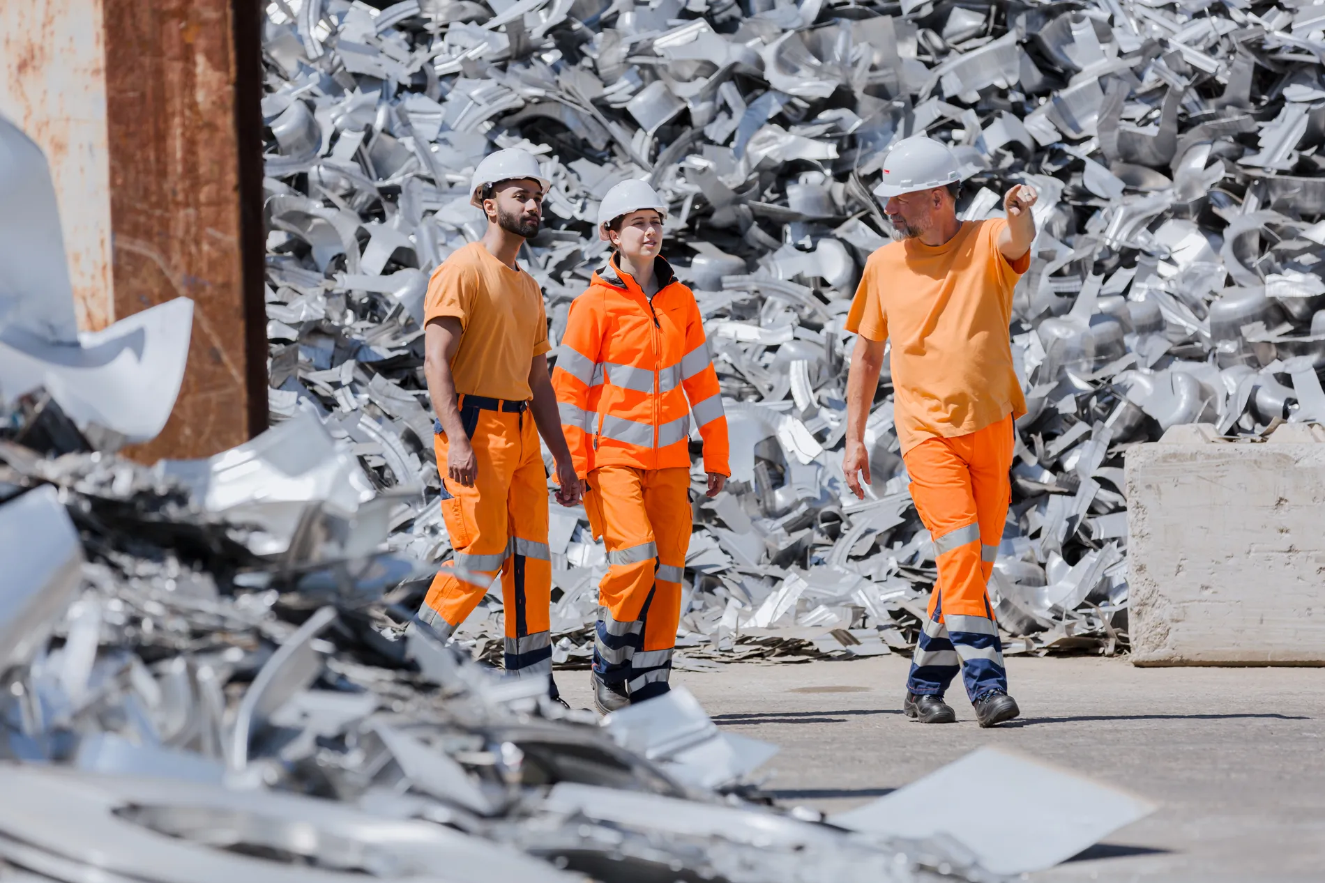 Drei Mitarbeitende in orangefarbener Arbeitskleidung und Helmen gehen gemeinsam über einen Recyclinghof mit Altmetallen.