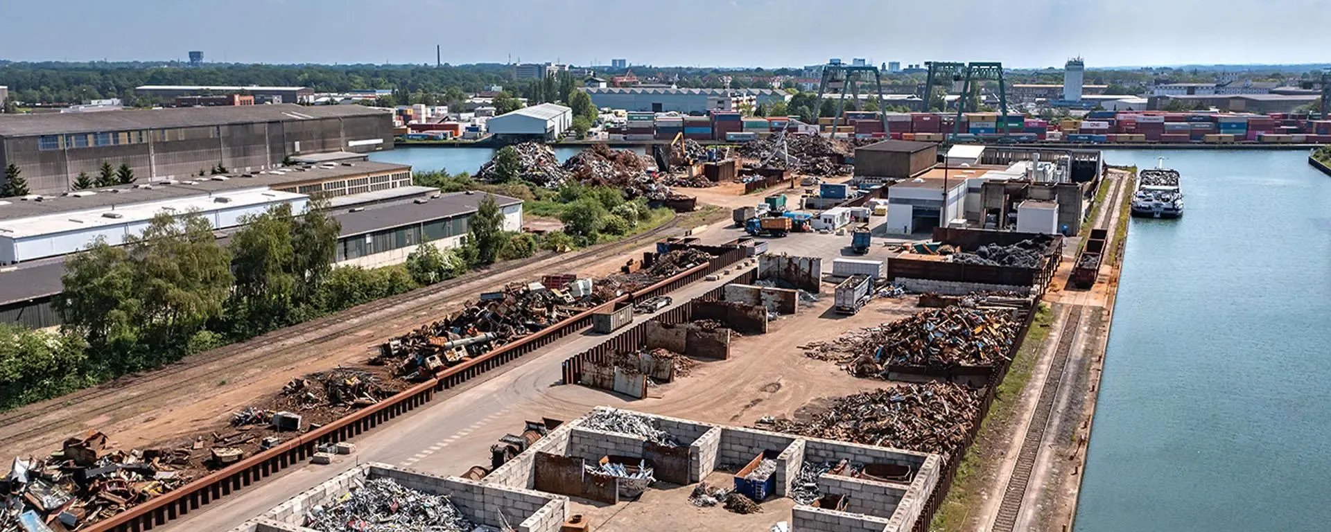 Luftaufnahme eines Recycling- und Umschlagplatzes von TSR Group am Wasser mit Containern und Schrott.