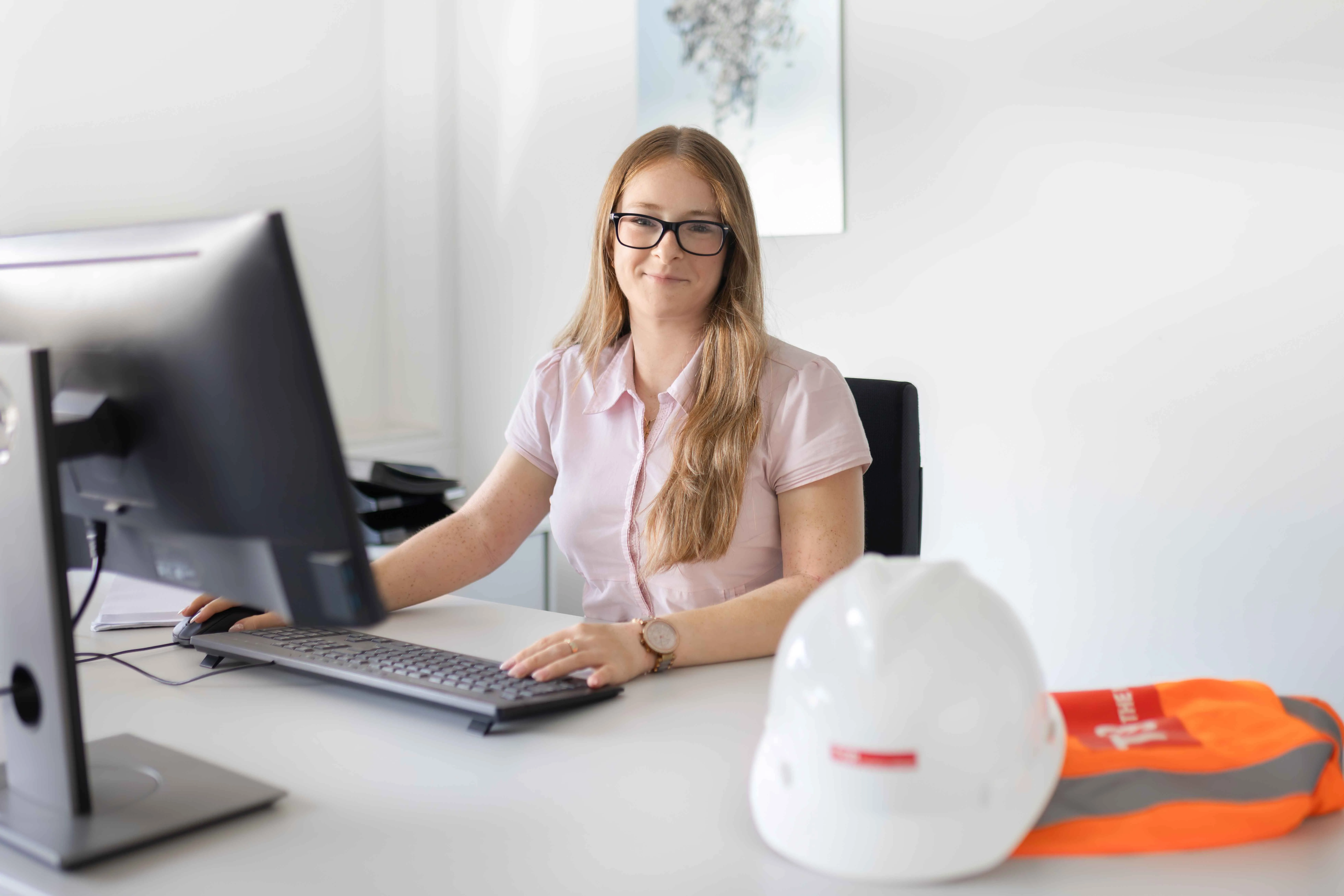 Junge Frau arbeitet am Schreibtisch im Büro, mit Schutzhelm und Warnweste von TSR Group im Vordergrund.