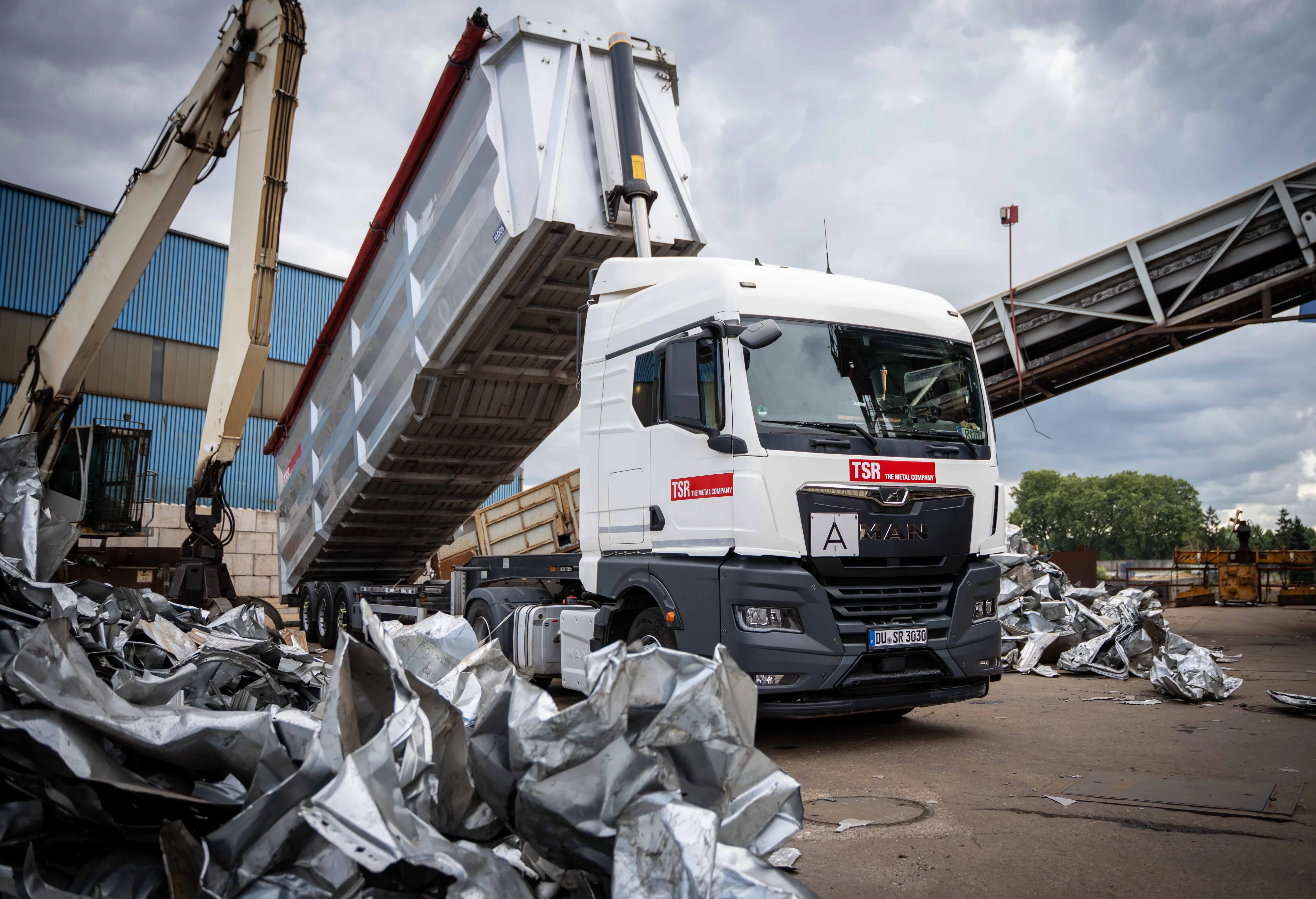 Drei Mitarbeitende in orangefarbener Arbeitskleidung und Helmen gehen gemeinsam über einen Recyclinghof mit Altmetallen.