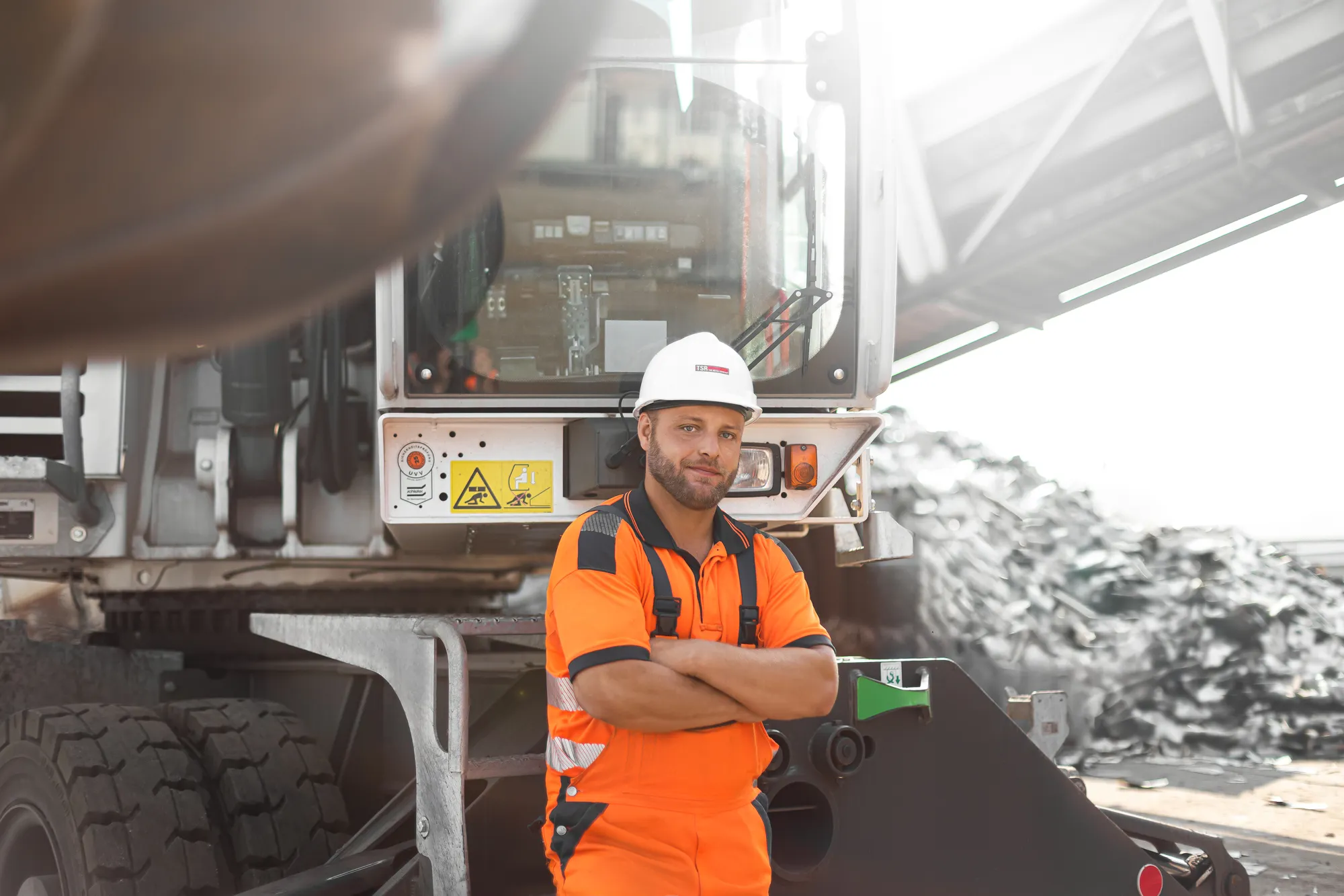 Ein Mitarbeiter in orangefarbener Arbeitskleidung und Helm steht vor einem Bagger auf dem TSR-Betriebsgelände.