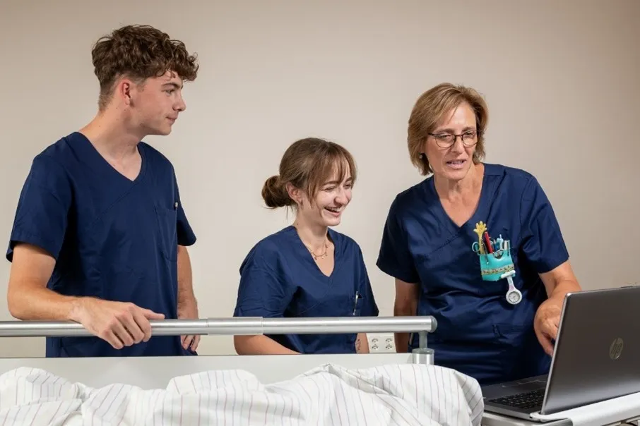 Drei Pflegekräfte im UKBB tauschen sich lächelnd vor einem Laptop am Krankenbett aus.