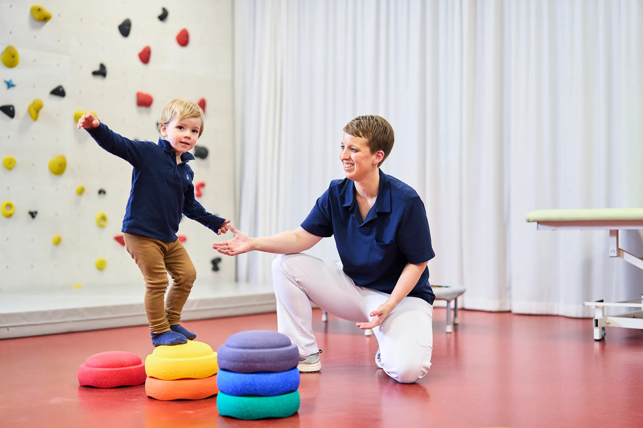Ein Kind balanciert auf einem Brett, unterstützt von einer Therapeutin im Rehabilitationsraum des UKBB.
