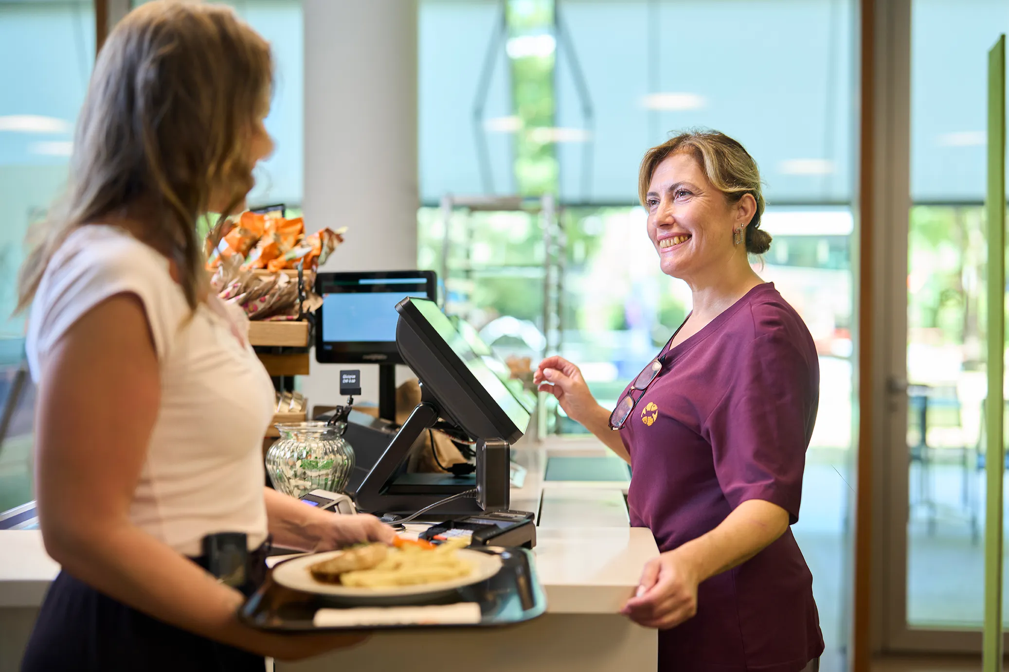 Mitarbeiterin der UKBB Cafeteria lächelt freundlich an der Kasse, während Du Dein Mittagessen bezahlst.