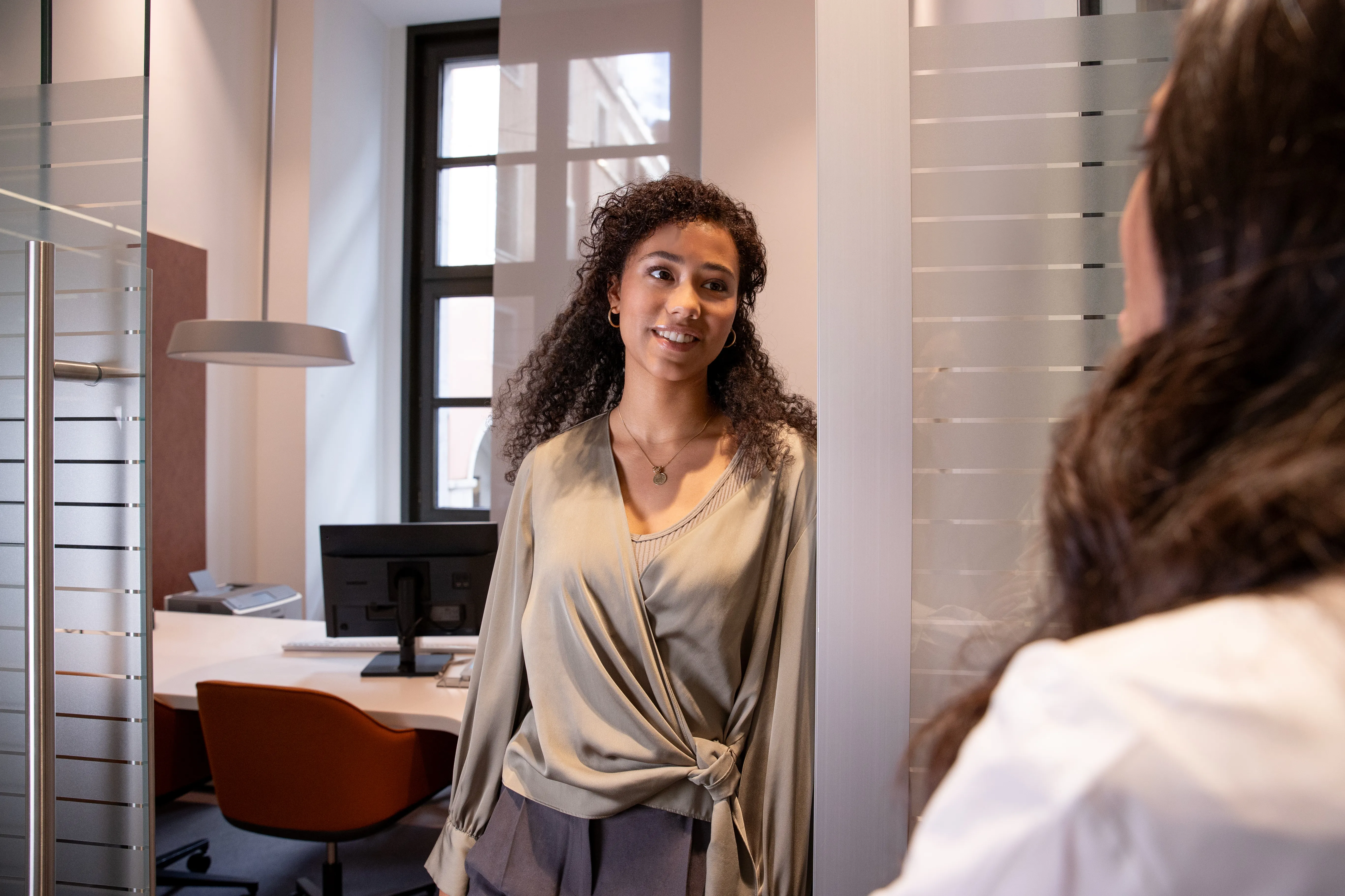 Eine junge Frau mit lockigem Haar lächelt in einem Büro und spricht mit einer Person im Vordergrund.