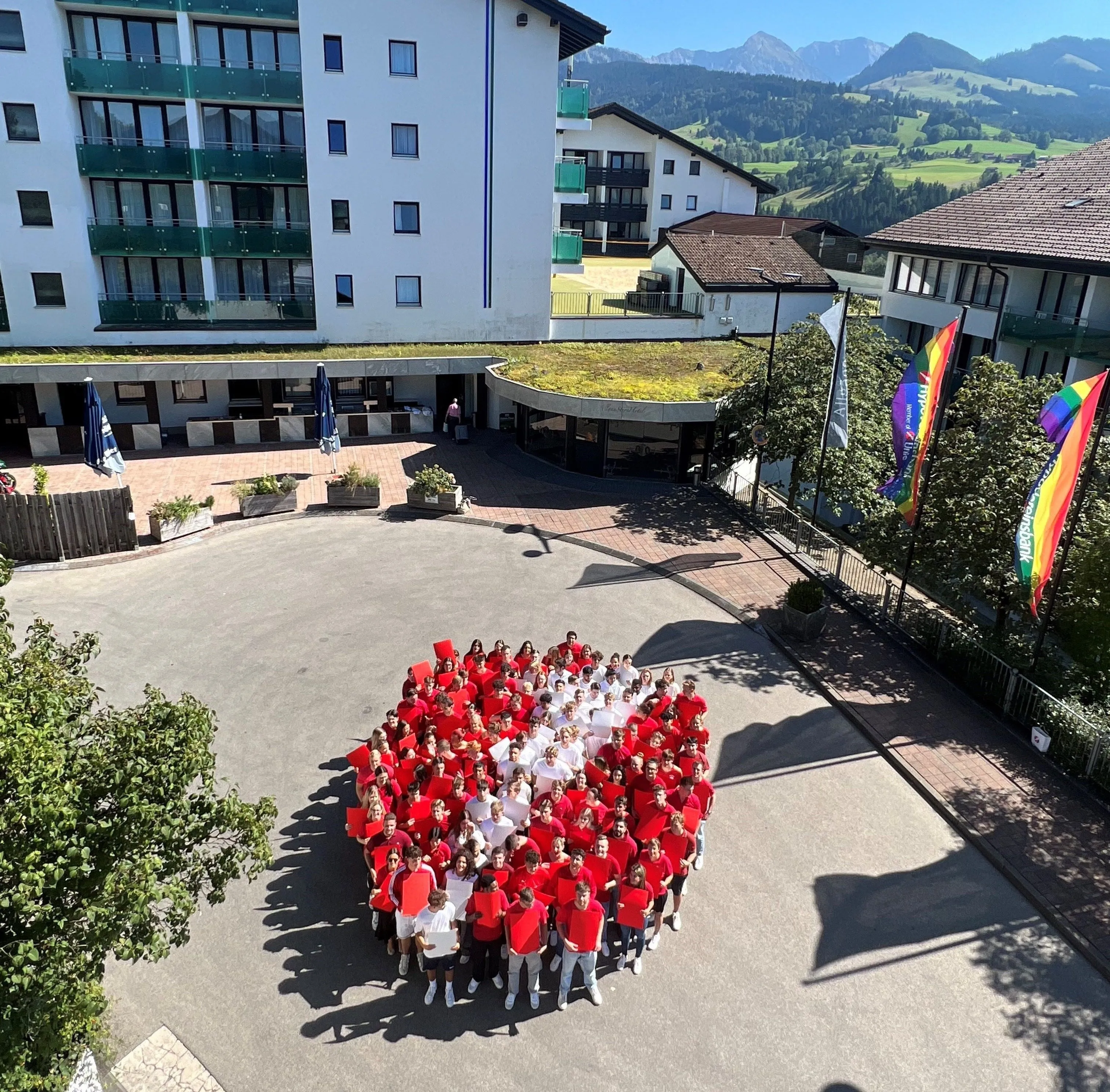 Eine Gruppe von Menschen formt die Zahl 1 in einem Innenhof, gekleidet in Rot und Weiß. Im Hintergrund sind Gebäude, Berge und Regenbogenfahnen sichtbar.