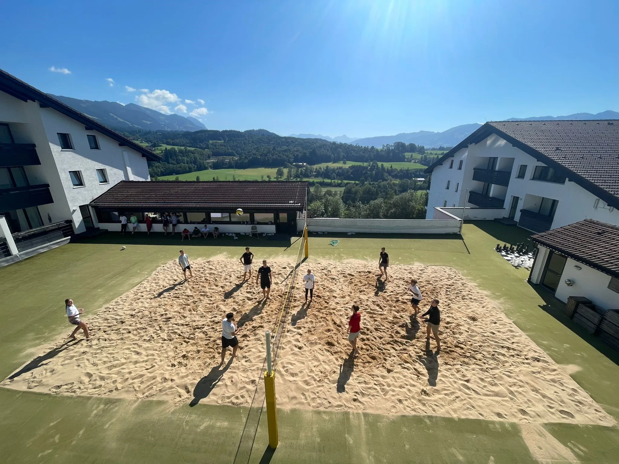 Mitarbeiter spielen Beachvolleyball auf einem Sandplatz vor einem Bürogebäude mit Bergblick.