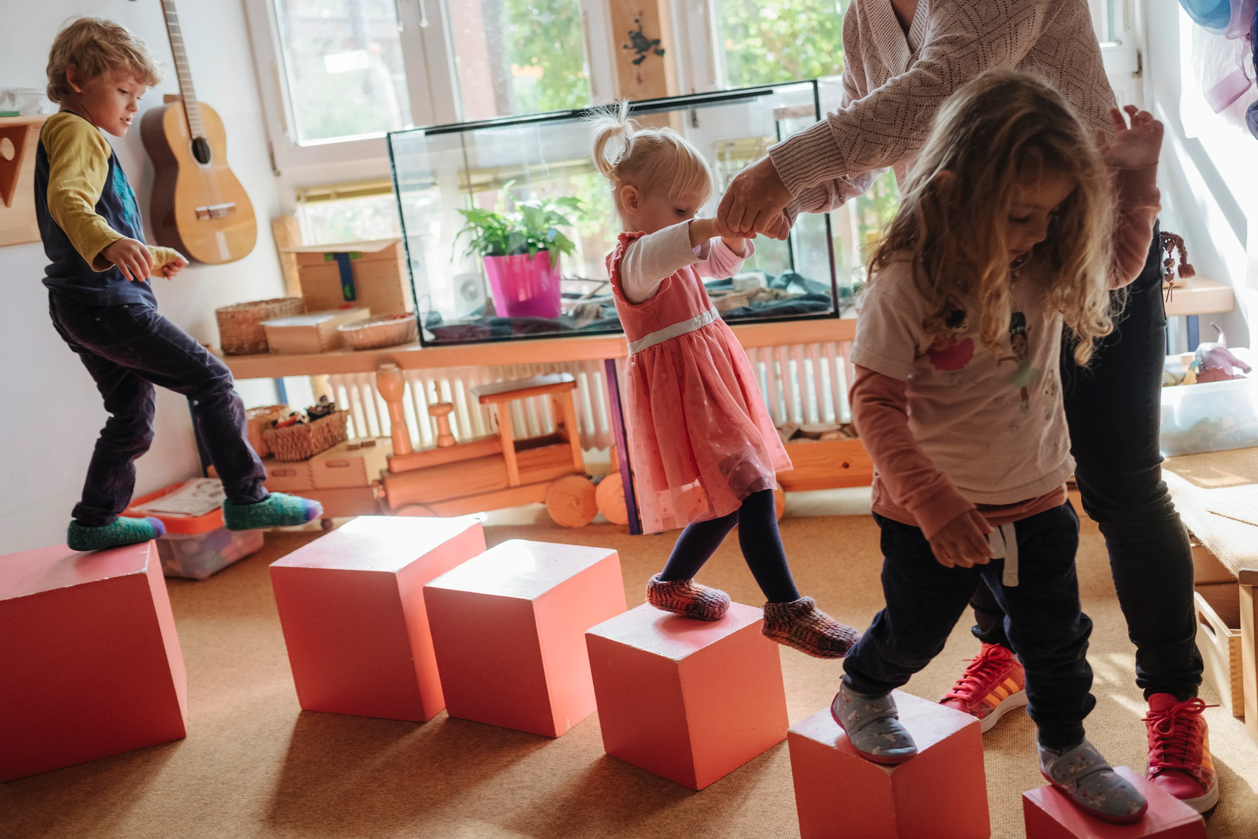 Kinder balancieren auf  Montessori-Würfeln in einer Kita, unterstützt von einer Erzieherin.