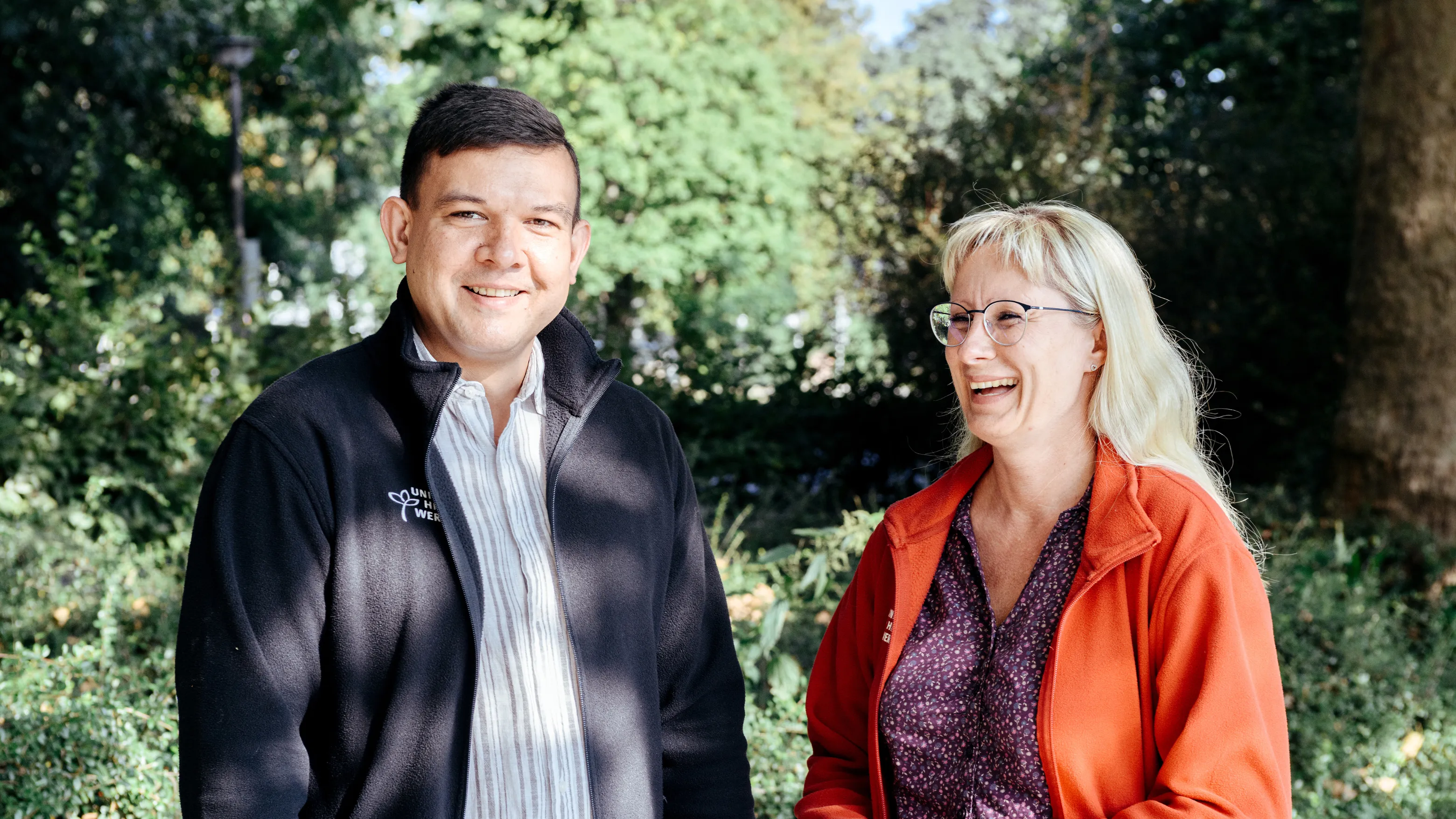 Zwei lächelnde Mitarbeiter des Unionhilfswerk im Gespräch im Freien, umgeben von grüner Natur.