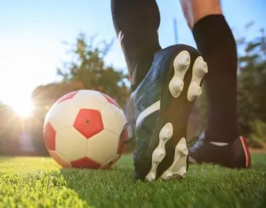 Nahaufnahme eines Fußballs und Fußballschuhen auf einem Spielfeld, mit Sonnenlicht im Hintergrund.