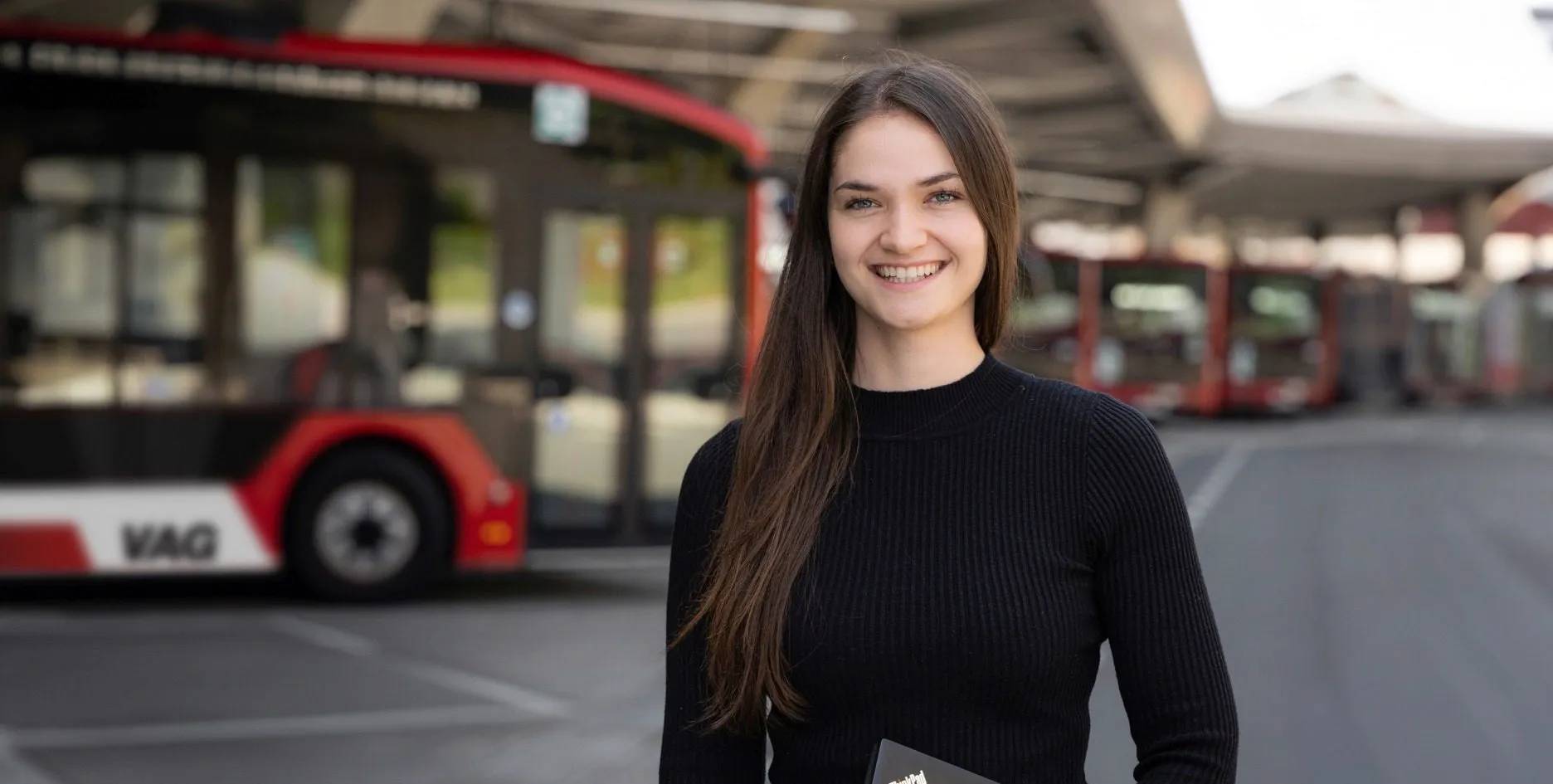 Junge Frau mit Laptop lächelt in moderner Werkstatt von VAG Verkehrs-Aktiengesellschaft, bereit für deine Karriere im Energie- und Nahverkehr.