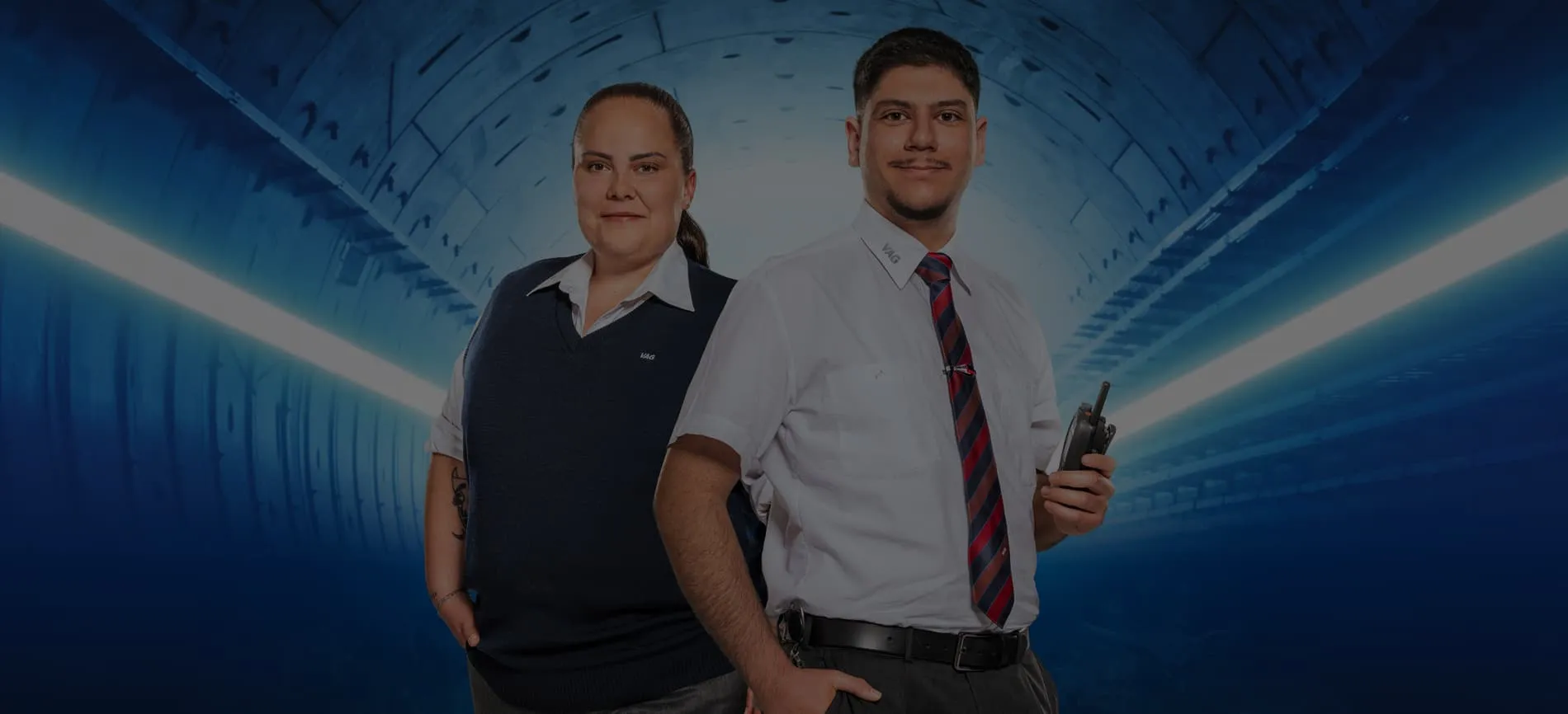 Zwei VAG-Mitarbeitende in Uniform stehen selbstbewusst in einem modernen Tunnel und blicken direkt in die Kamera.