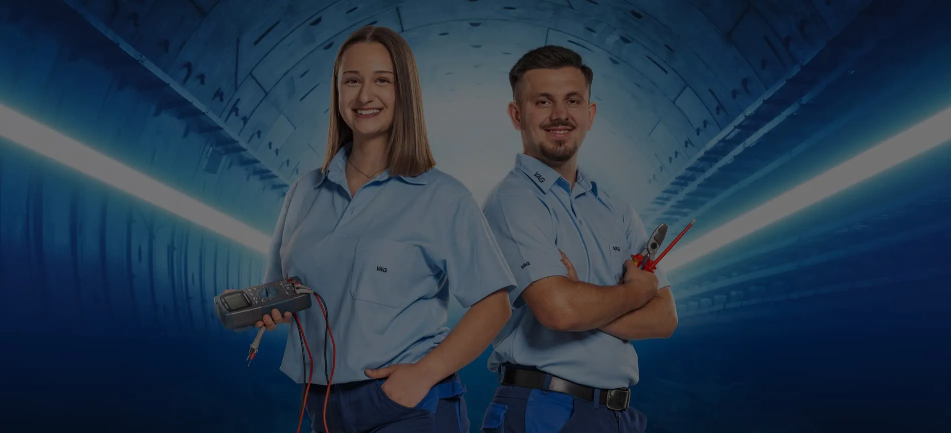 Zwei junge VAG-Mitarbeitende in Arbeitskleidung mit Werkzeug im Tunnel, lächeln und wirken einladend.