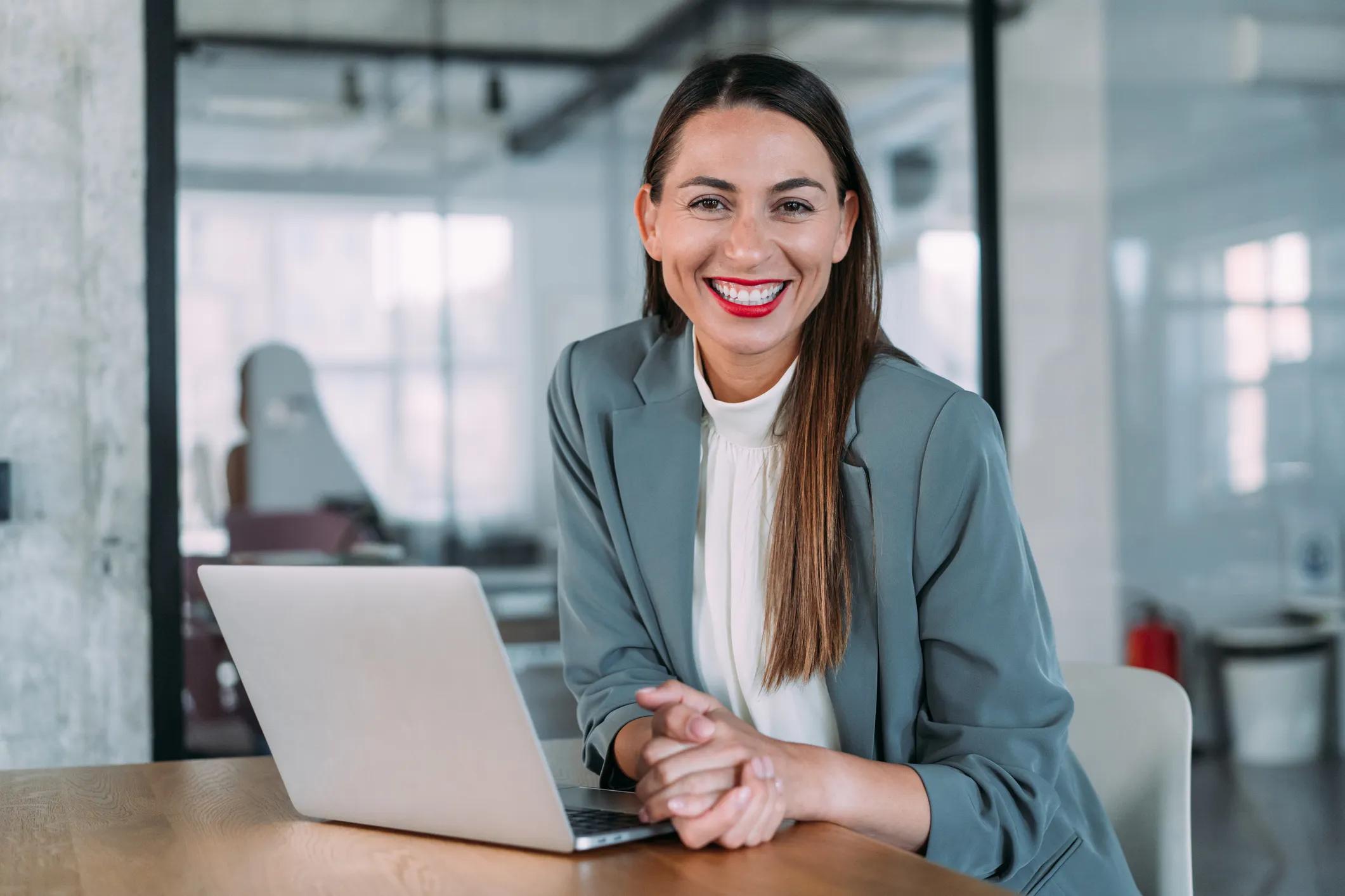 Junge Bankmitarbeiterin im Business-Outfit lächelt Dich am Schreibtisch mit Laptop im modernen Büro an.