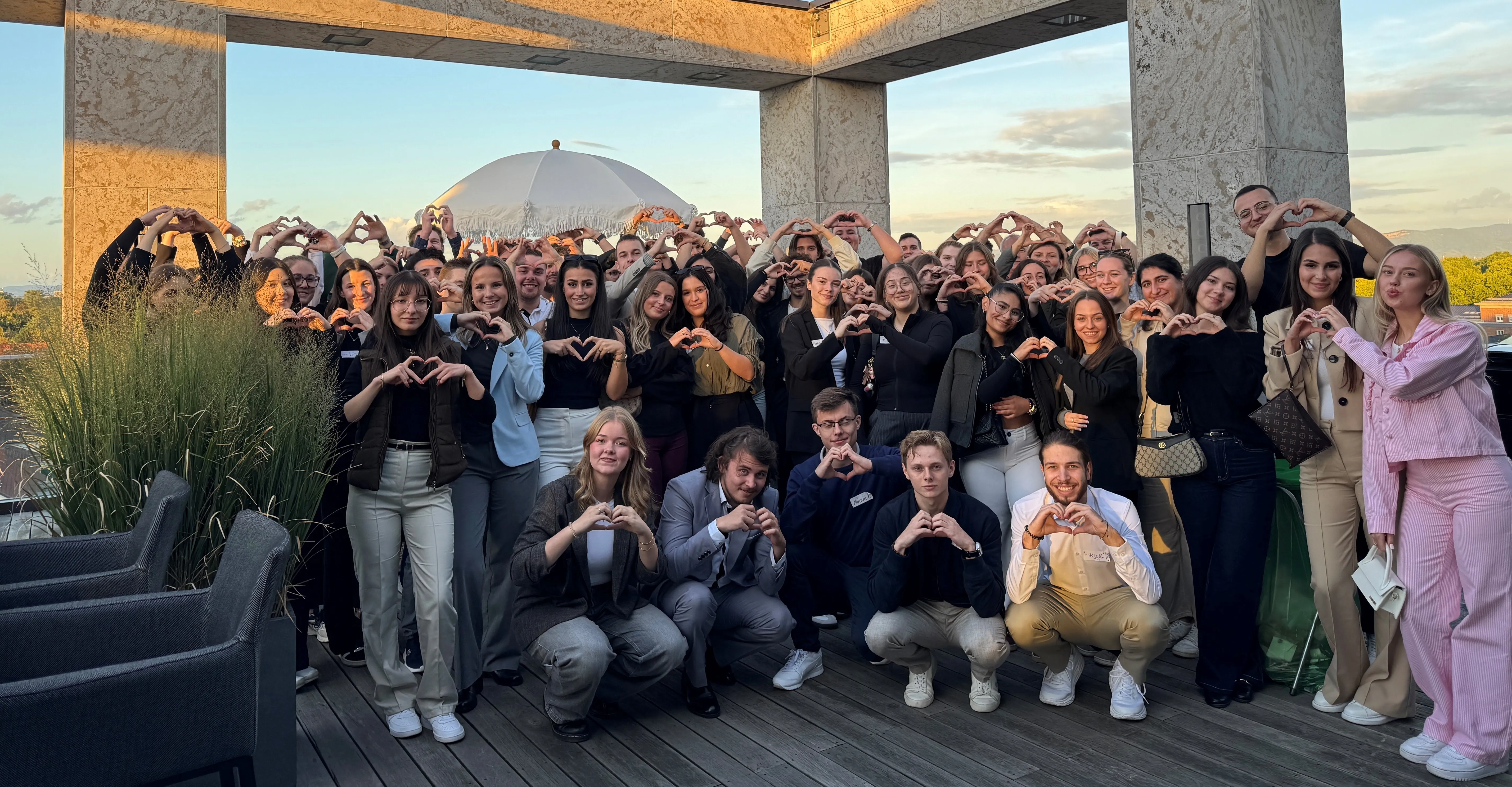 Junges Team der VR Bank steht lächelnd zusammen auf einer Terrasse und formt mit den Händen Herzzeichen.