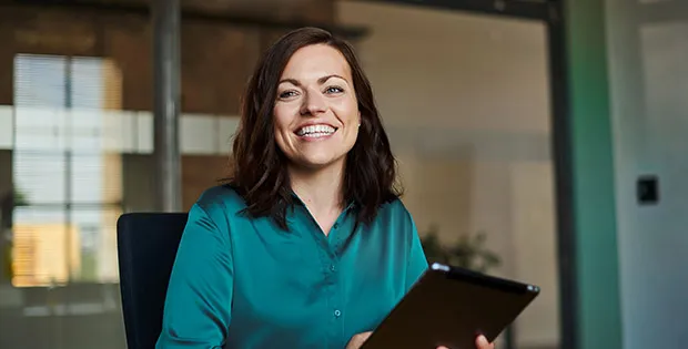 Junge Frau im Büro der VR Bank lächelt dich an und hält ein Tablet, bereit für neue Karrieremöglichkeiten.
