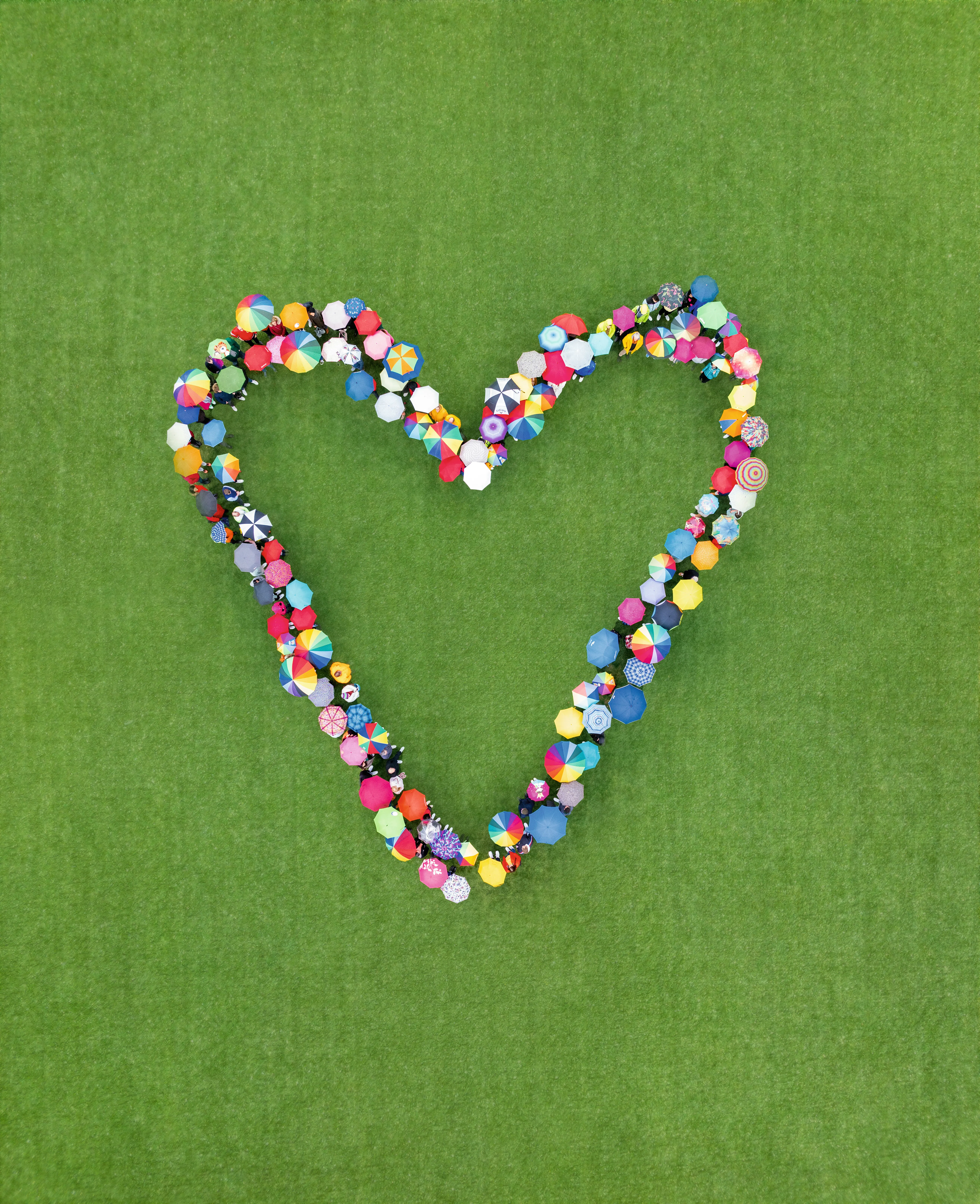 Viele Menschen mit bunten Regenschirmen bilden gemeinsam ein Herz auf einer grünen Wiese – Teamgeist bei VR Bank.