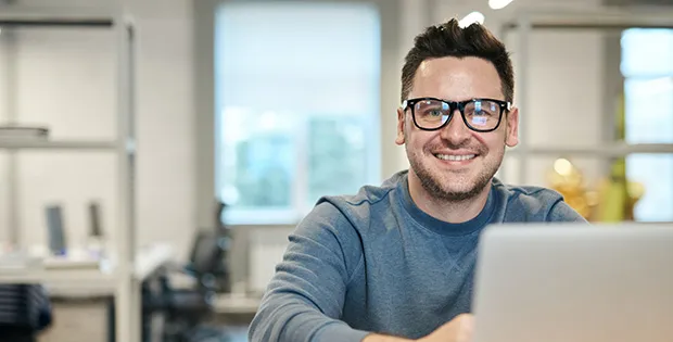 Junger Mann mit Brille arbeitet lächelnd an einem Laptop in einem modernen Büro der VR Bank.