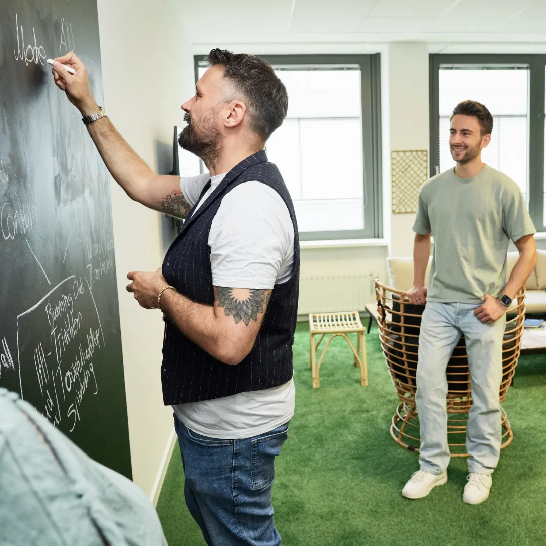 Ein Mann im orangefarbenen Pullover betrachtet aufmerksam eine Pinnwand im Büro. Im Vordergrund sind unscharfe Personen zu sehen.
