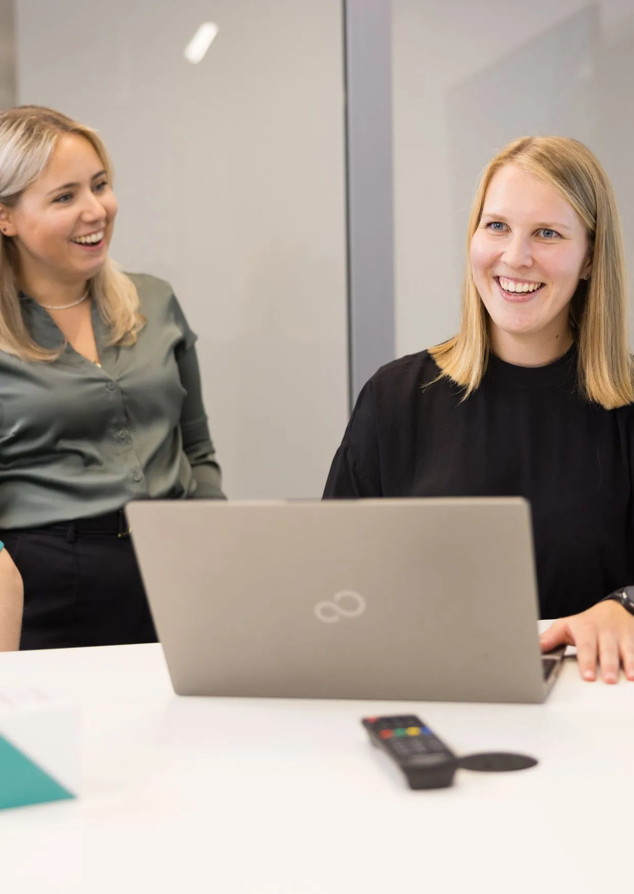 Zwei junge Frauen lachen gemeinsam am Arbeitsplatz, eine sitzt am Laptop, die andere steht daneben im modernen Büro.