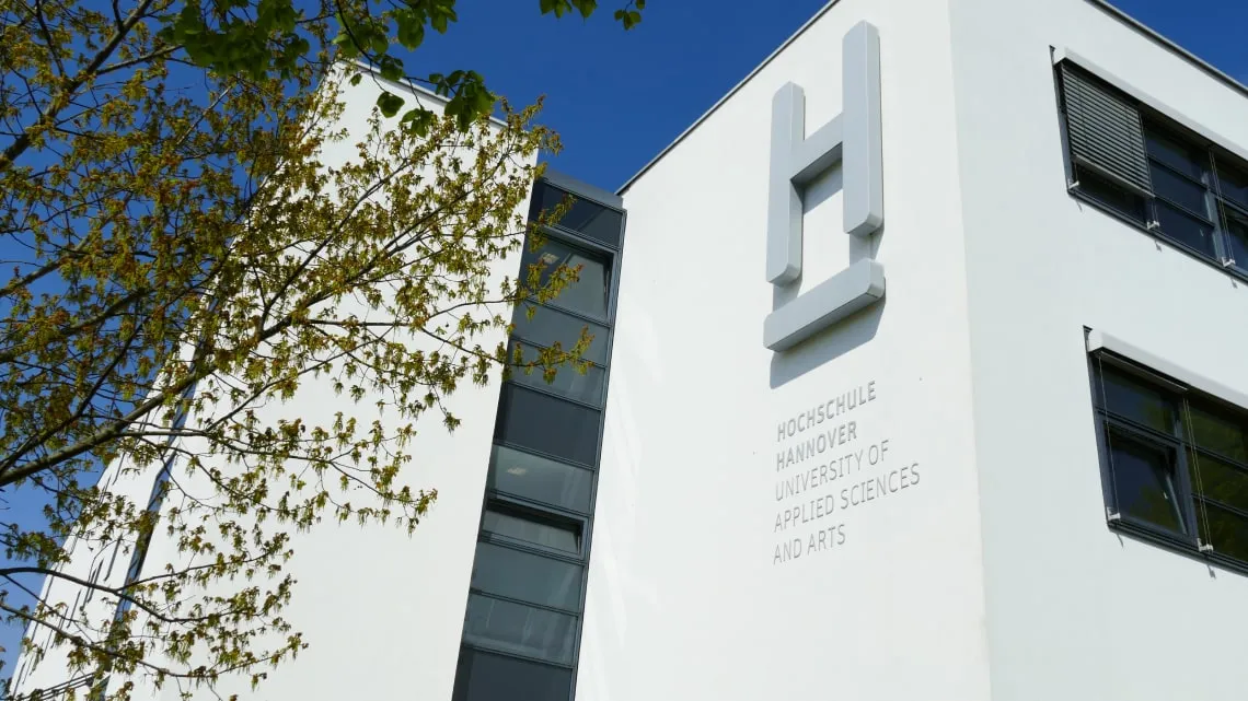 Modernes Hochschulgebäude mit Logo und Schriftzug „Hochschule Hannover“, umgeben von grünen Bäumen im Frühling.