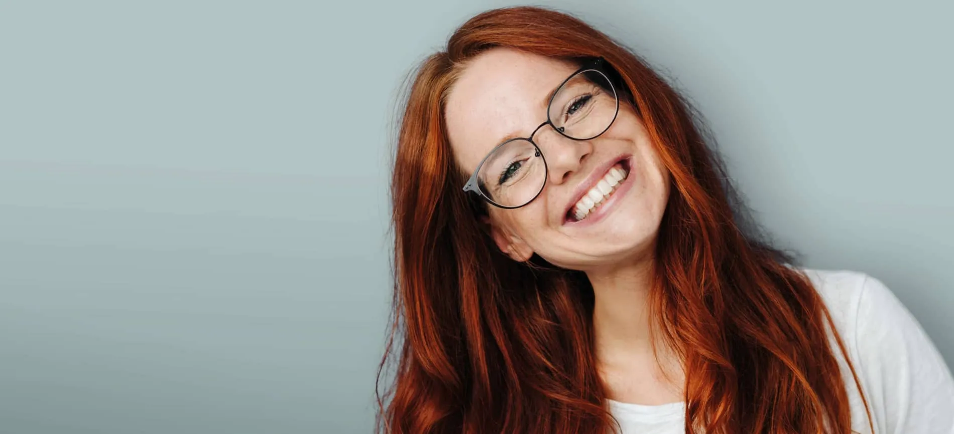 Junge Frau mit langen roten Haaren und Brille lächelt dich freundlich vor neutralem Hintergrund an.