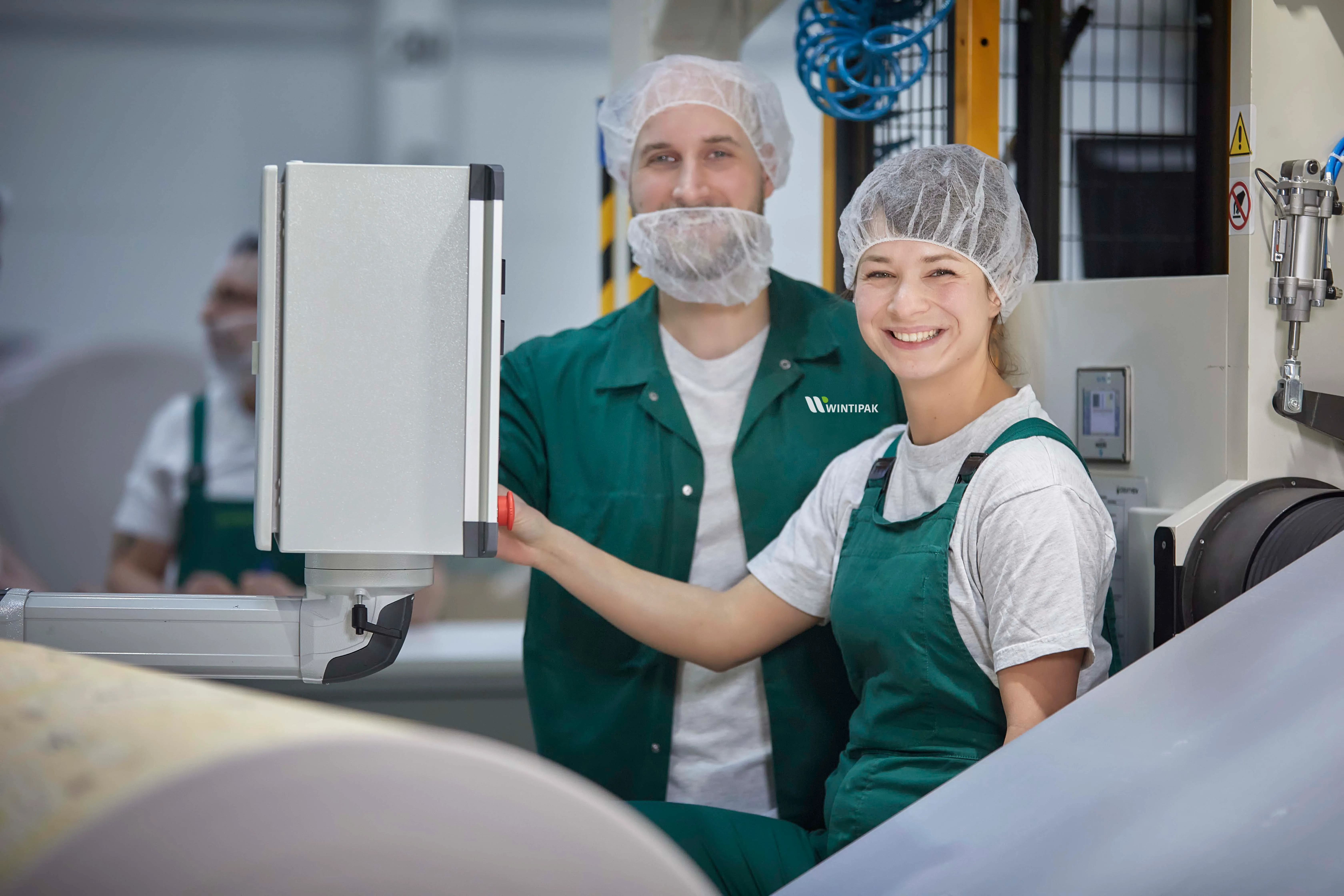 Zwei Mitarbeiter von Wintipak in grüner Arbeitskleidung und Haarnetzen bedienen eine Maschine und lächeln in die Kamera.