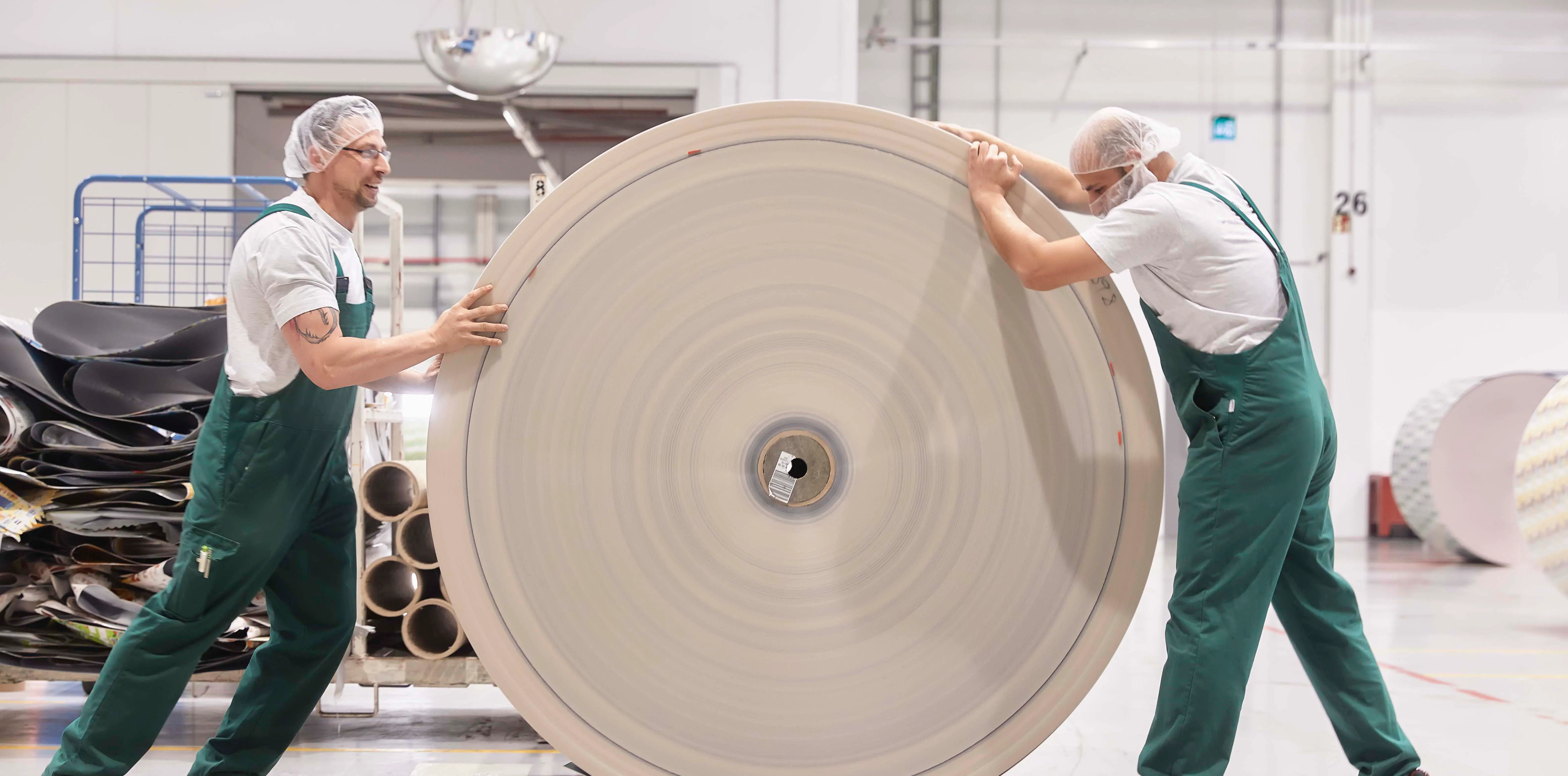 Zwei Mitarbeiter in grünen Overalls rollen eine große Papierrolle in einer Produktionshalle von Wintipak.