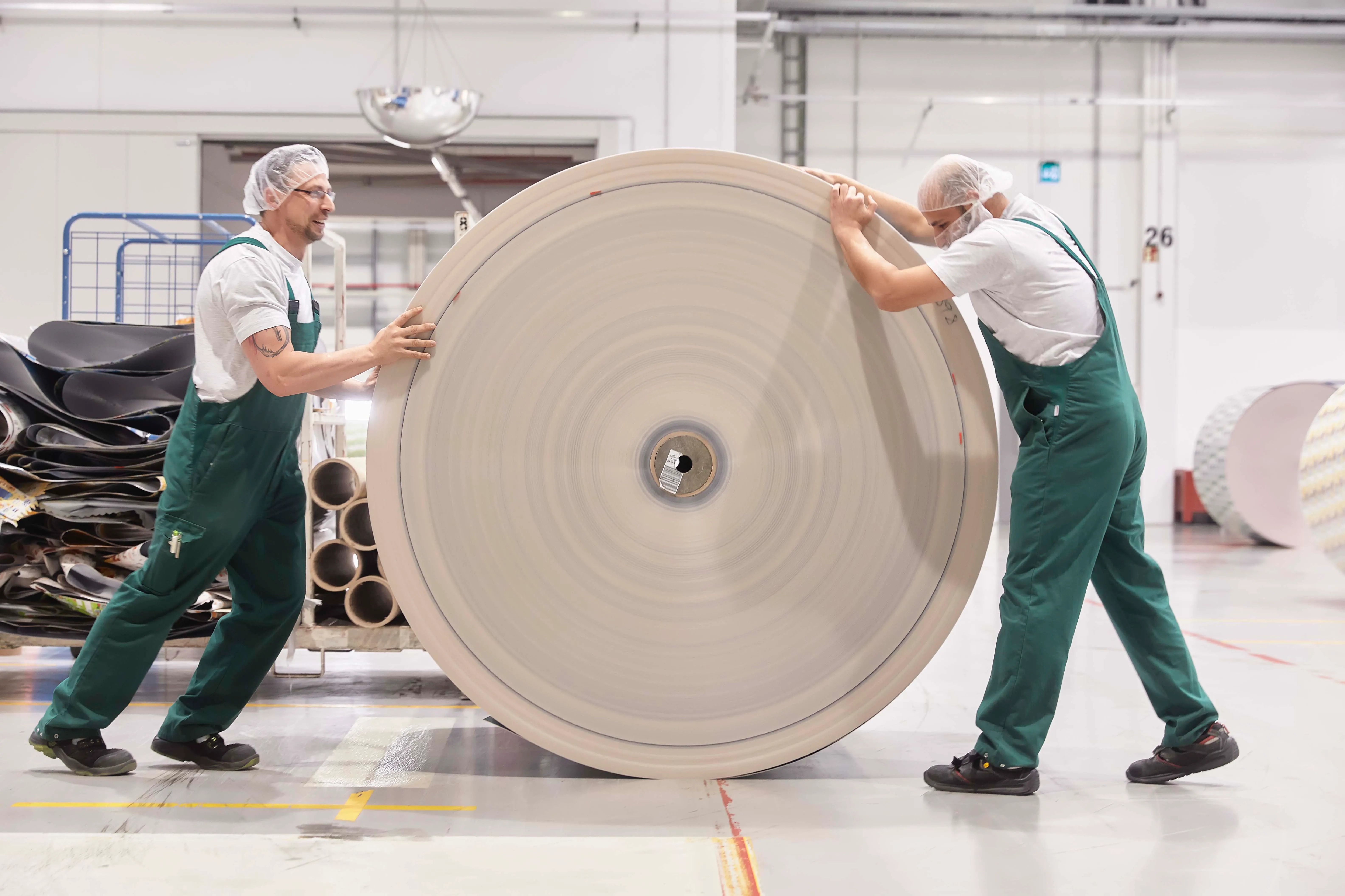 Zwei Arbeiter in grünen Overalls bewegen eine große Papierrolle in einer Fabrikhalle von Wintipak.
