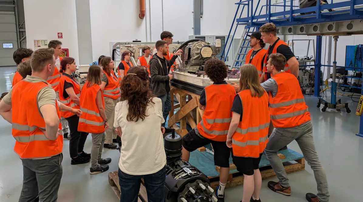 Mehrere junge Menschen in orangefarbenen Warnwesten bekommen bei ZF eine Erklärung zu einem Automobilbauteil in der Werkshalle.