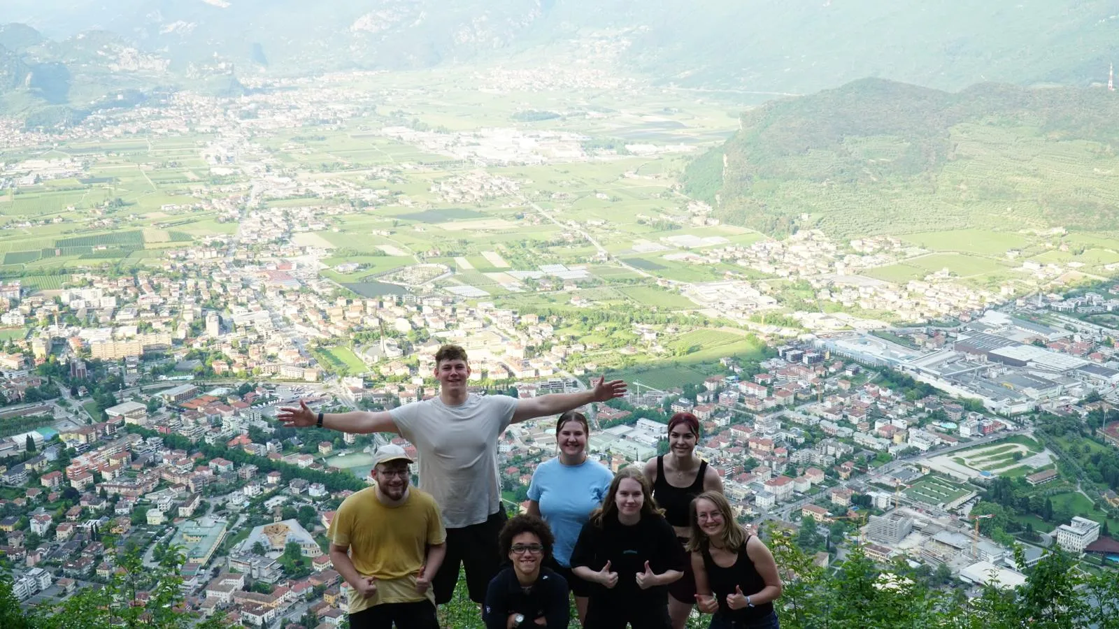 Eine Gruppe junger ZF-Mitarbeitender posiert fröhlich auf einem Berg mit weitem Blick über eine Stadtlandschaft.