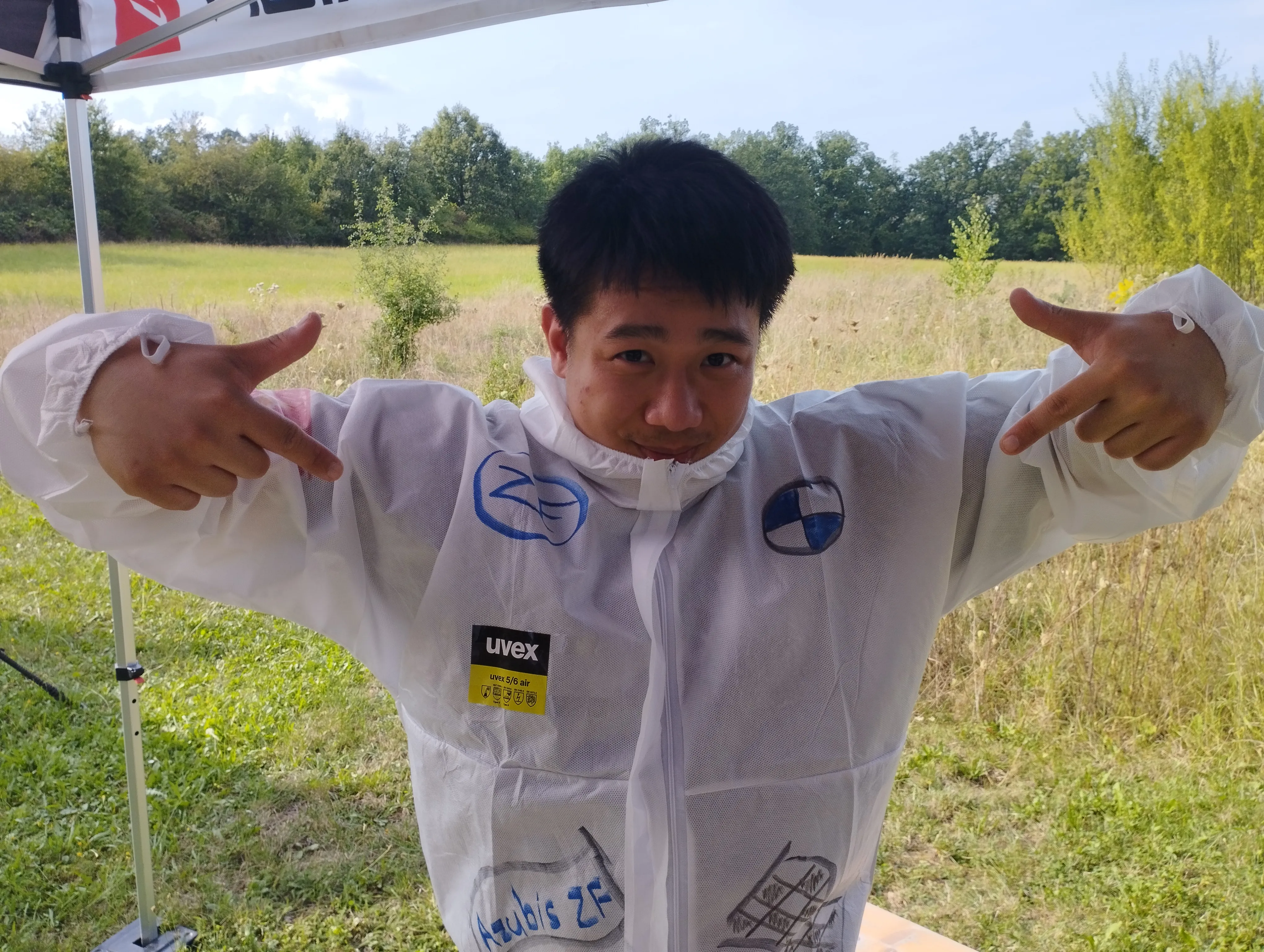 Junger Mann im Schutzanzug mit ZF-Logo zeigt lachend auf „Azubis ZF“ – entdecke Deine Ausbildung bei ZF im Automotive-Bereich.