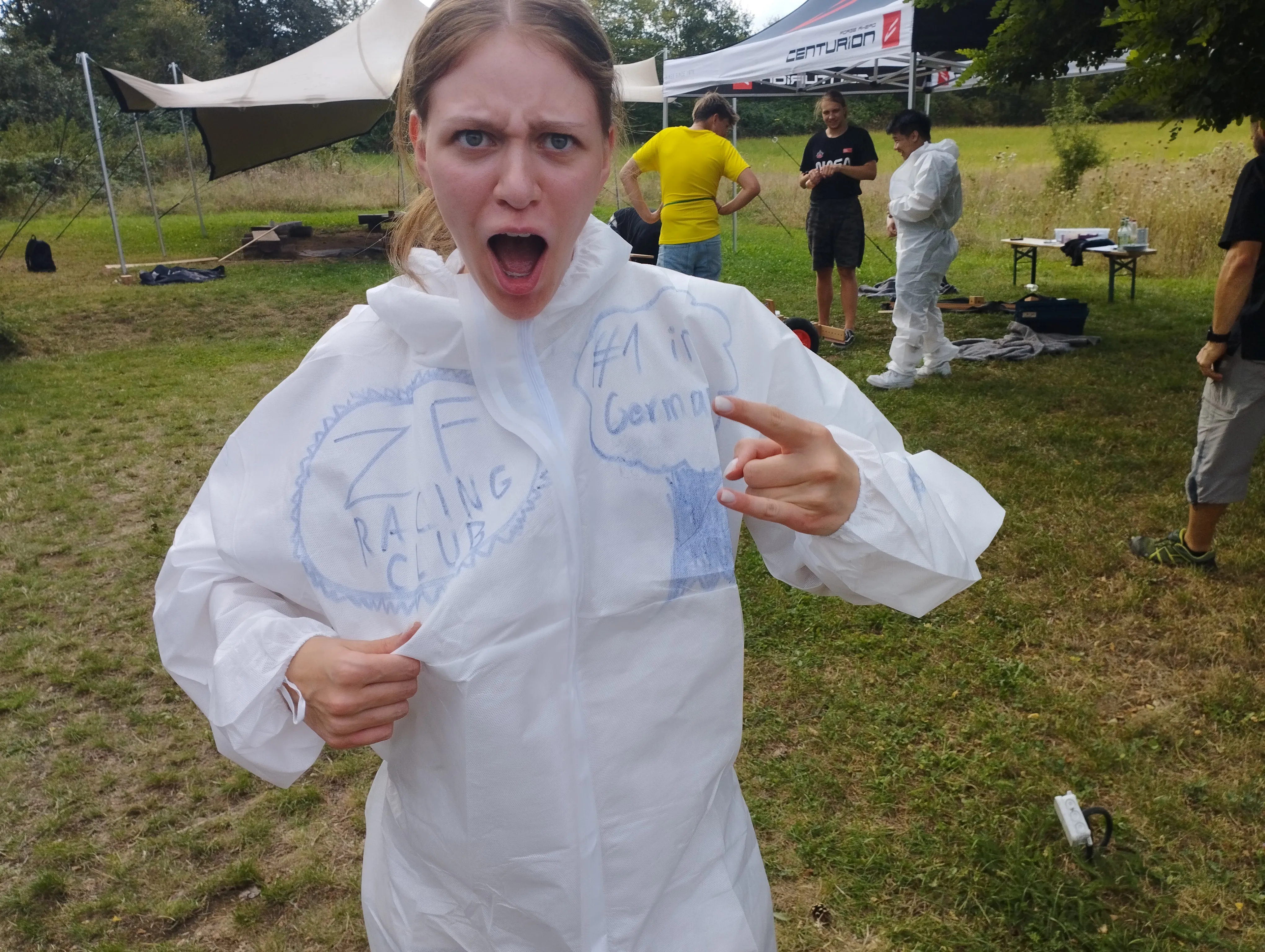Junge Frau im weißen Overall mit „ZF Racing Club“-Aufschrift posiert motiviert beim Outdoor-Teamevent von ZF Friedrichshafen AG.