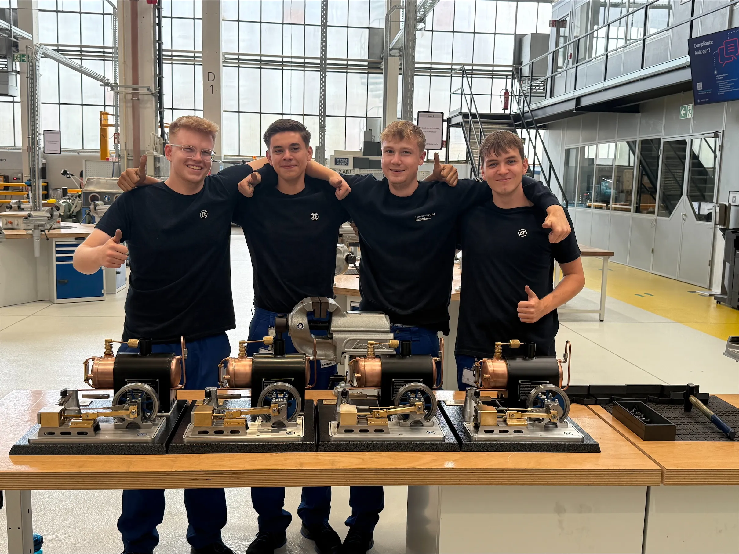 Vier junge Auszubildende bei ZF stehen lachend in der Werkstatt hinter selbstgebauten Modellmotoren.